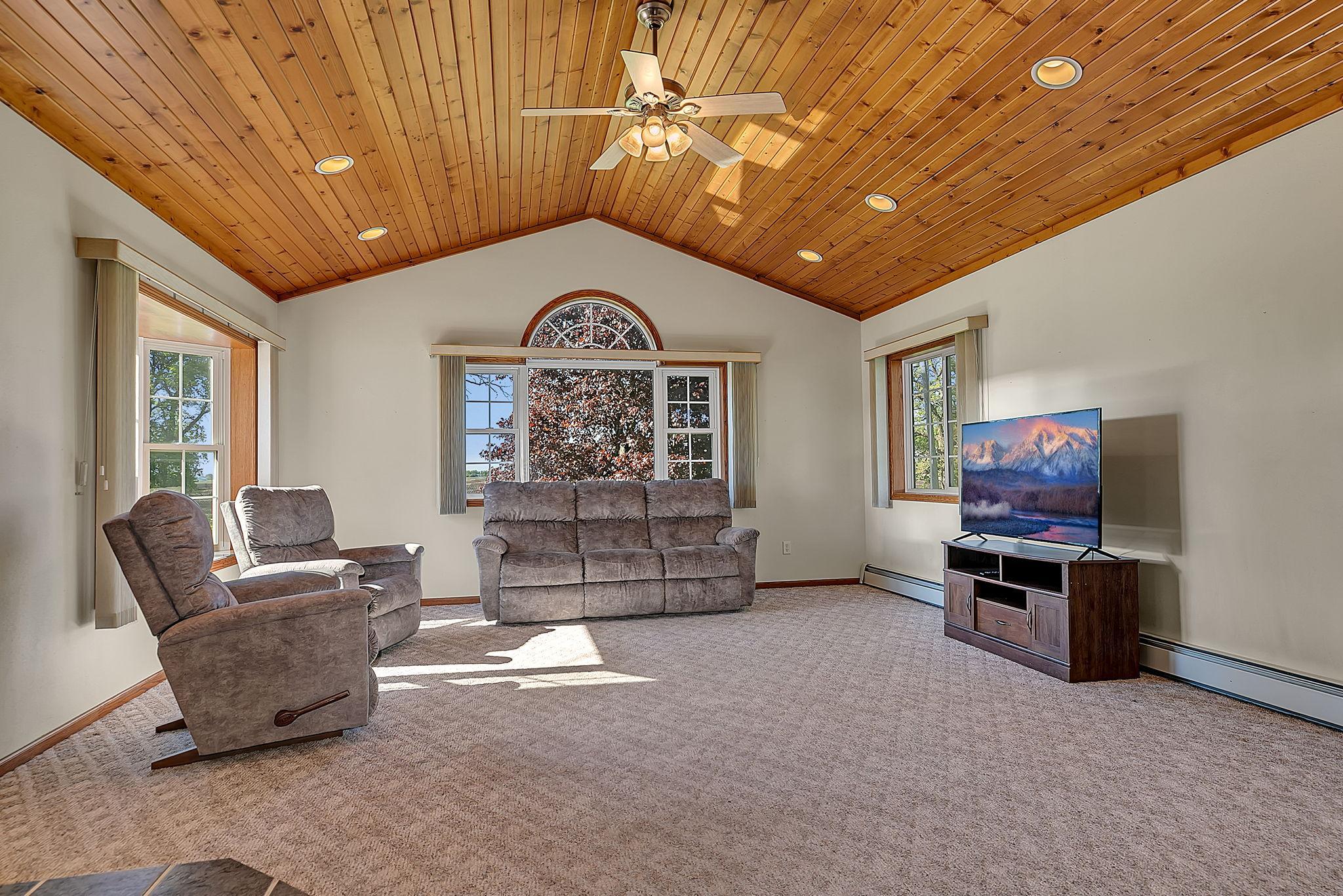 Spacious living room showcasing a vaulted wood ceiling with recessed lighting and ceiling fan, creating a warm and inviting atmosphere. Large windows fill the space with natural light. Vitually staged