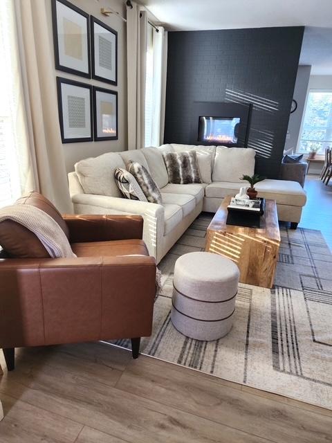 Sun-drenched and stunning — the living room features wide plank luxury vinyl flooring, oversized windows, and a double-sided gas fireplace with a dramatic black brick facade.