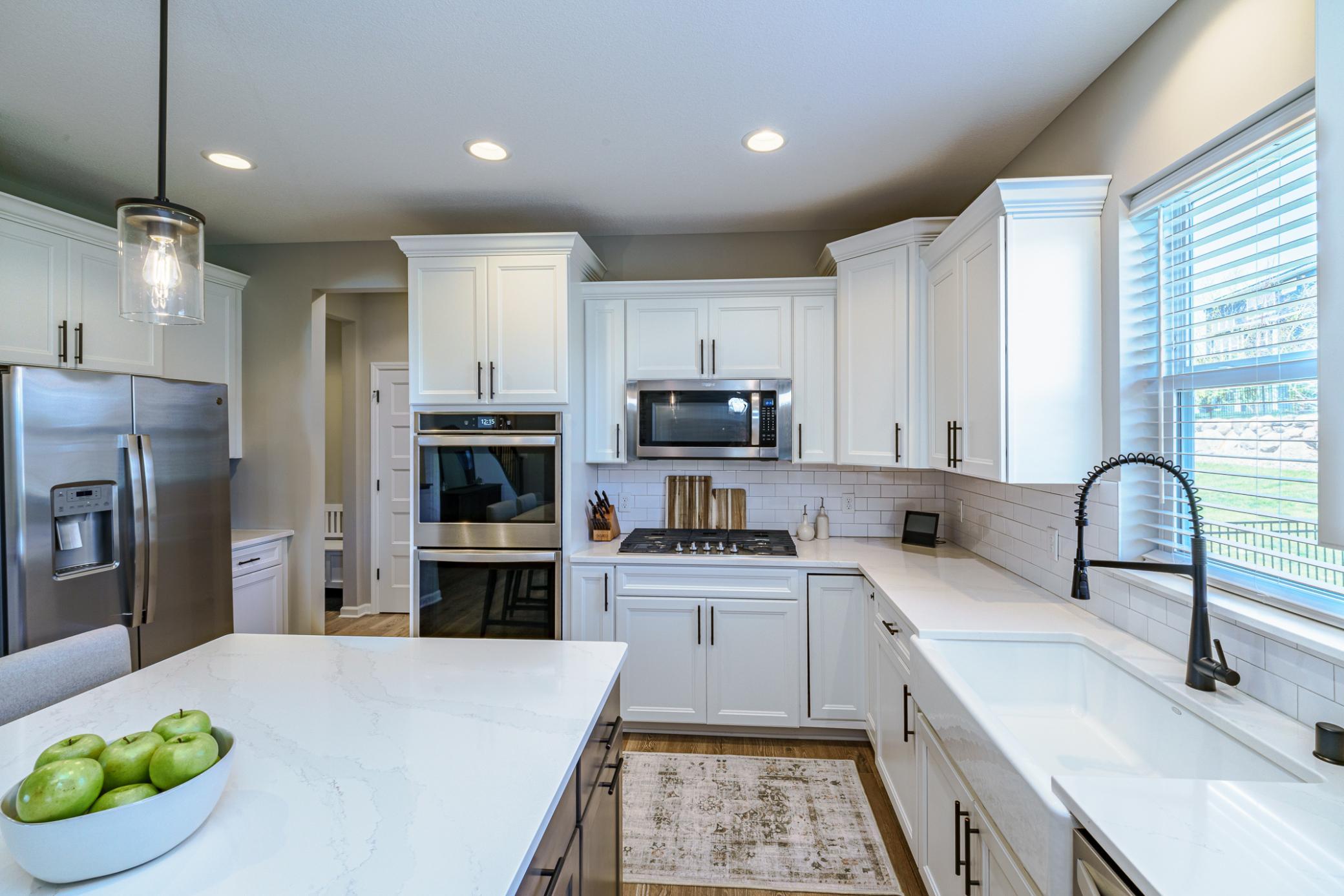A subway tile backsplash offers classic balance to the quartz countertops. The kitchen has a farmhouse sink with a gooseneck faucet underneath the bright window, as well as stainless appliances including a gas cooktop and a double oven.