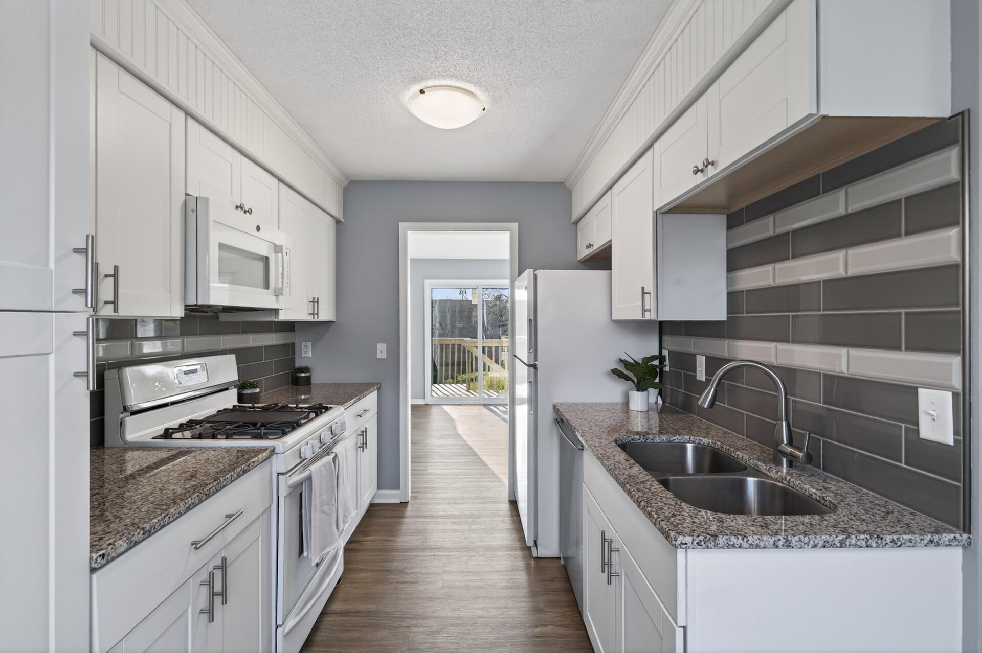Bright, updated galley kitchen featuring white cabinetry, granite countertops, subway tile backsplash, stainless sink, and direct sightline to the deck and outdoor space.