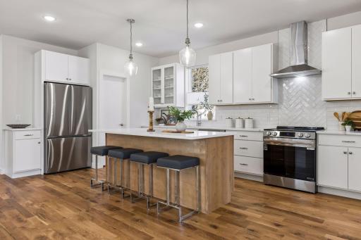Chef-inspired kitchen with quartz countertops, large center island with seating and abundant cabinetry offering plenty of shelving. Features a kitchen window for natural light, modern hood, pantry storage and stainless steel appliances.
