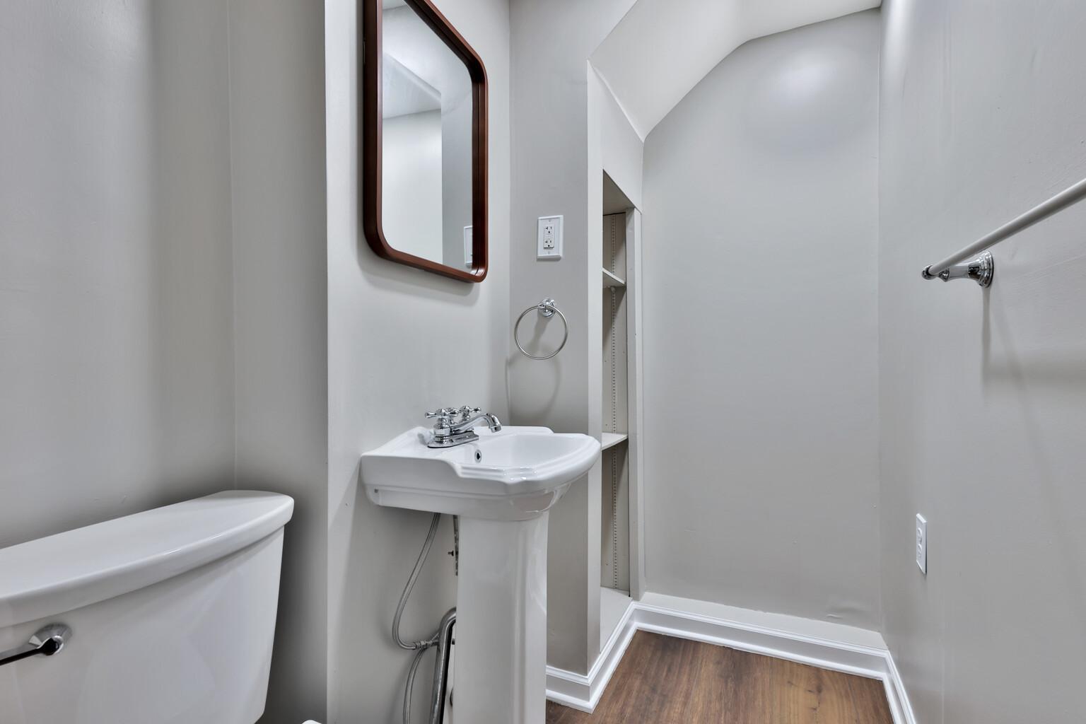 Bright lower-level half bath featuring a classic pedestal sink. Includes integrated recessed shelving for easy storage and modern wood-look flooring. A clean, polished, and comfortable addition.
