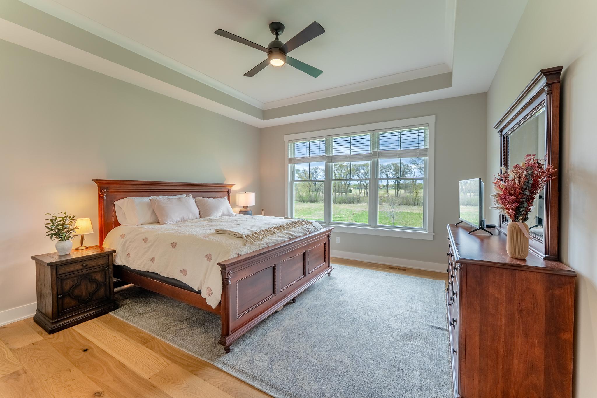 A tranquil primary suite with a tray ceiling, ceiling fan, rich hardwood furniture tones, and sweeping nature views that make waking up feel like a getaway.