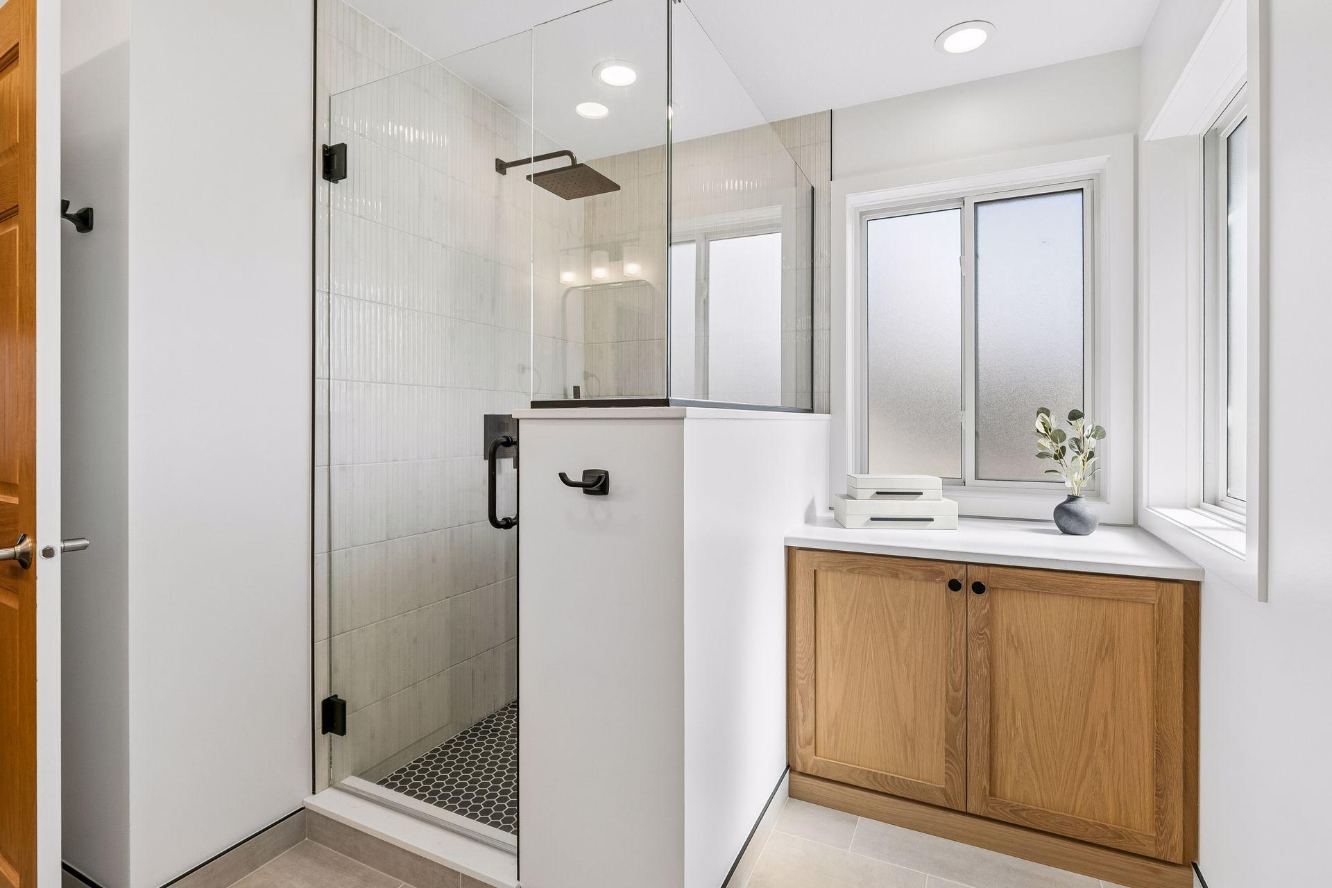 This view of the primary bath showcases a walk-in tiled shower with frameless glass. A bright, clean, and tasteful design. An added bonus of this remodel is the built-in white oak cabinet and new corner windows with privacy glass.