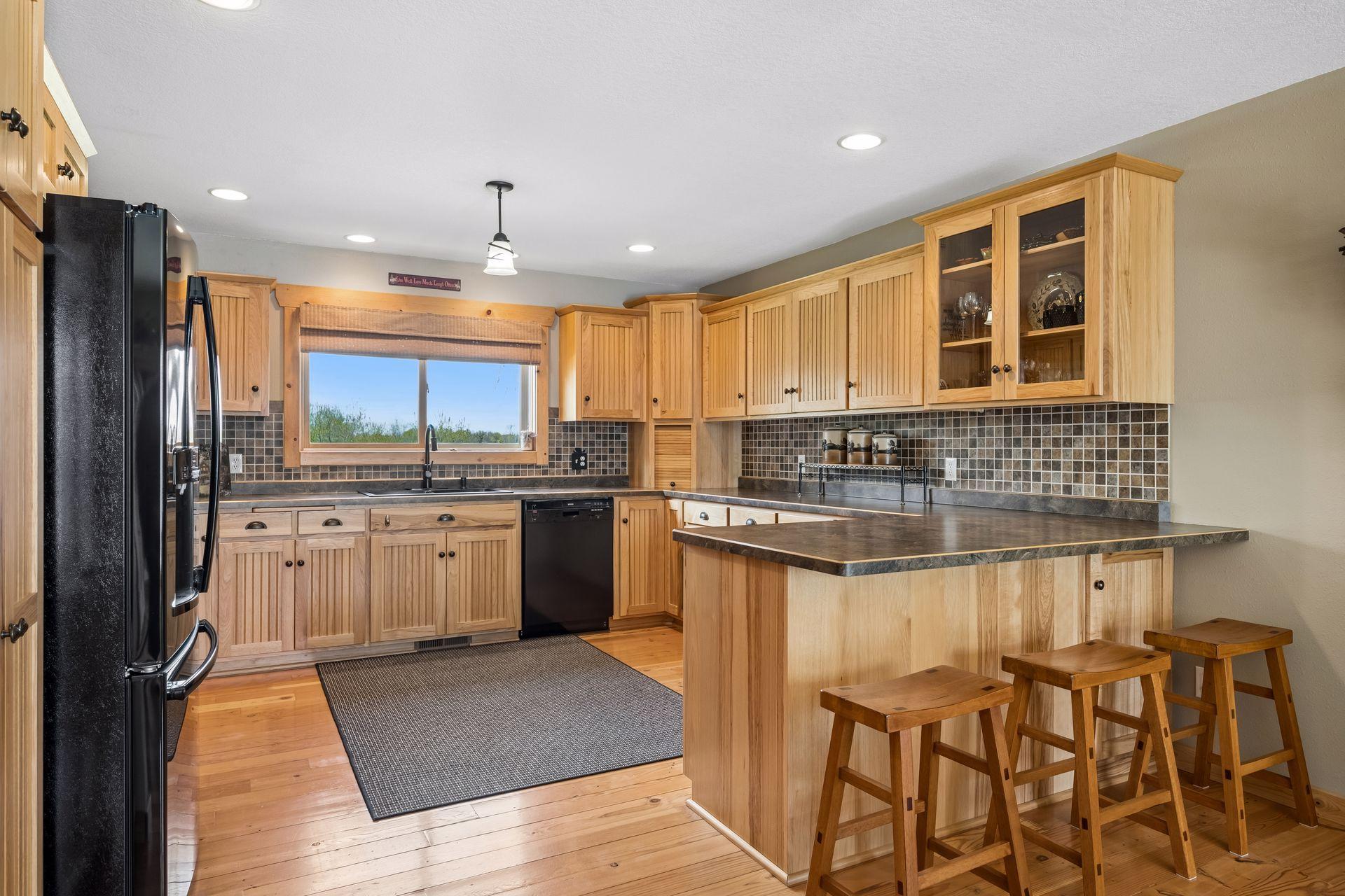 Warm and inviting kitchen showcasing beautiful hickory cabinetry, ample storage, and natural wood finishes. Functional layout with breakfast bar seating, tile backsplash, and large window bringing in light—perfect for everyday living.