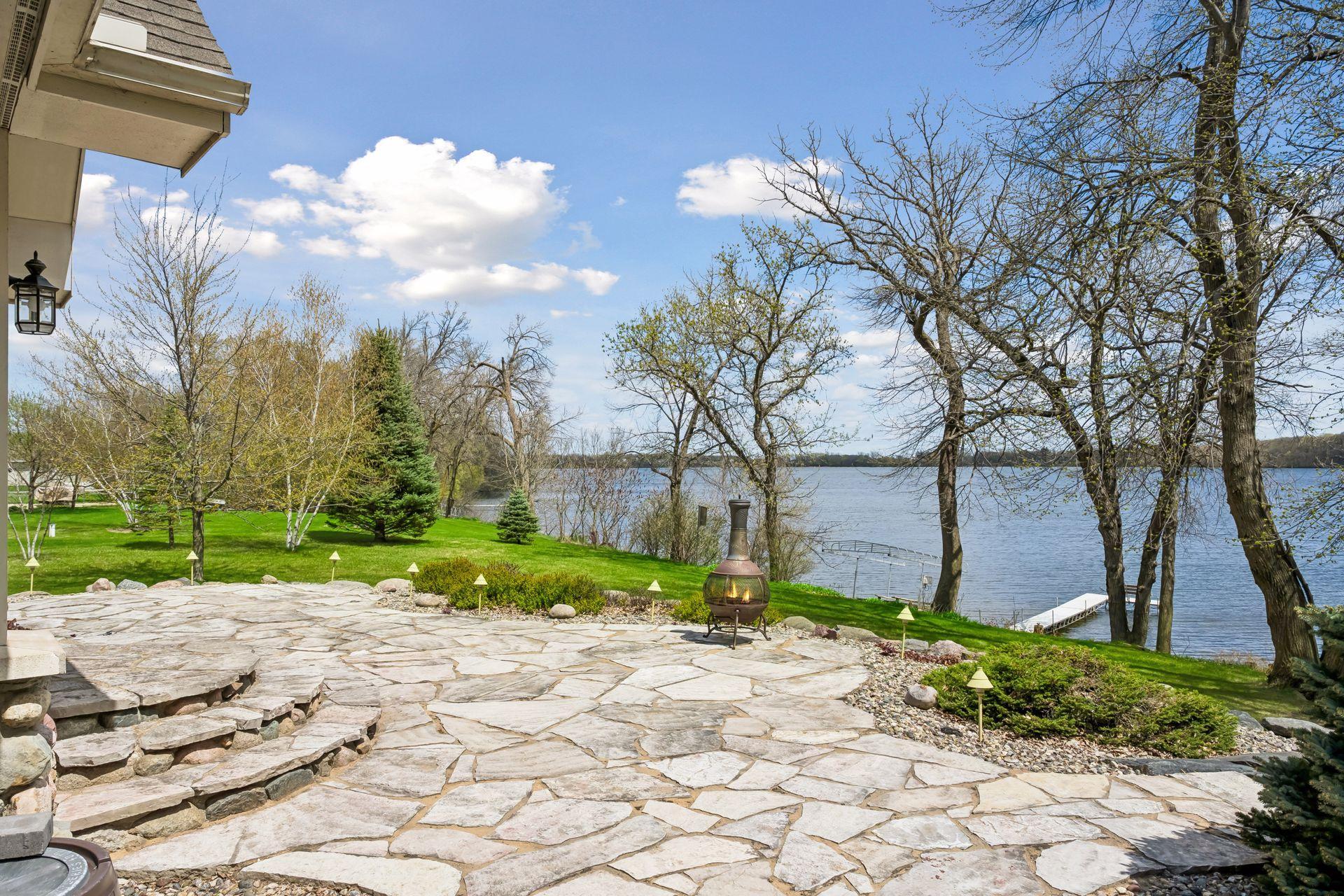 Beautiful lakeside setting on a full recreational lake in Dassel, featuring a spacious flagstone patio, mature trees, manicured lawn, and private dock. Enjoy peaceful water views, outdoor fire feature, and ideal space for relaxing or entertaining