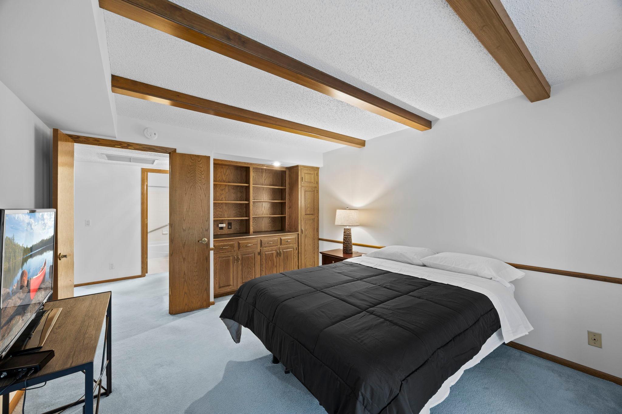 The second bedroom is equally generous in scale and versatility, ideal as a bedroom, home office, or additional living space. It includes plush carpeting, a full wall of built-in wood cabinetry and shelving, and charming wood beam ceiling accents.