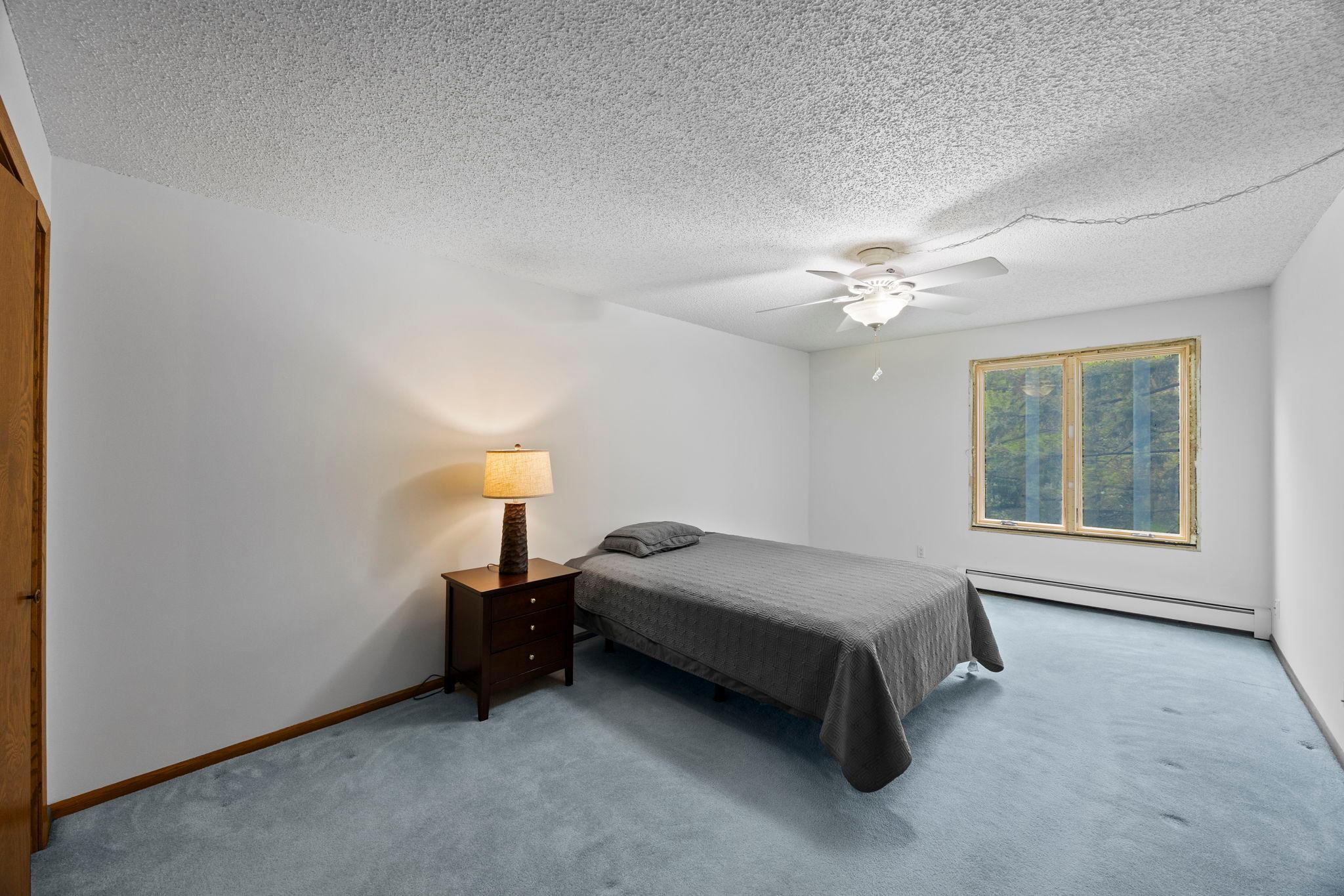 The spacious primary suite features soft carpeting, a ceiling fan, a large window with lush greenery views, and an oversized closet.