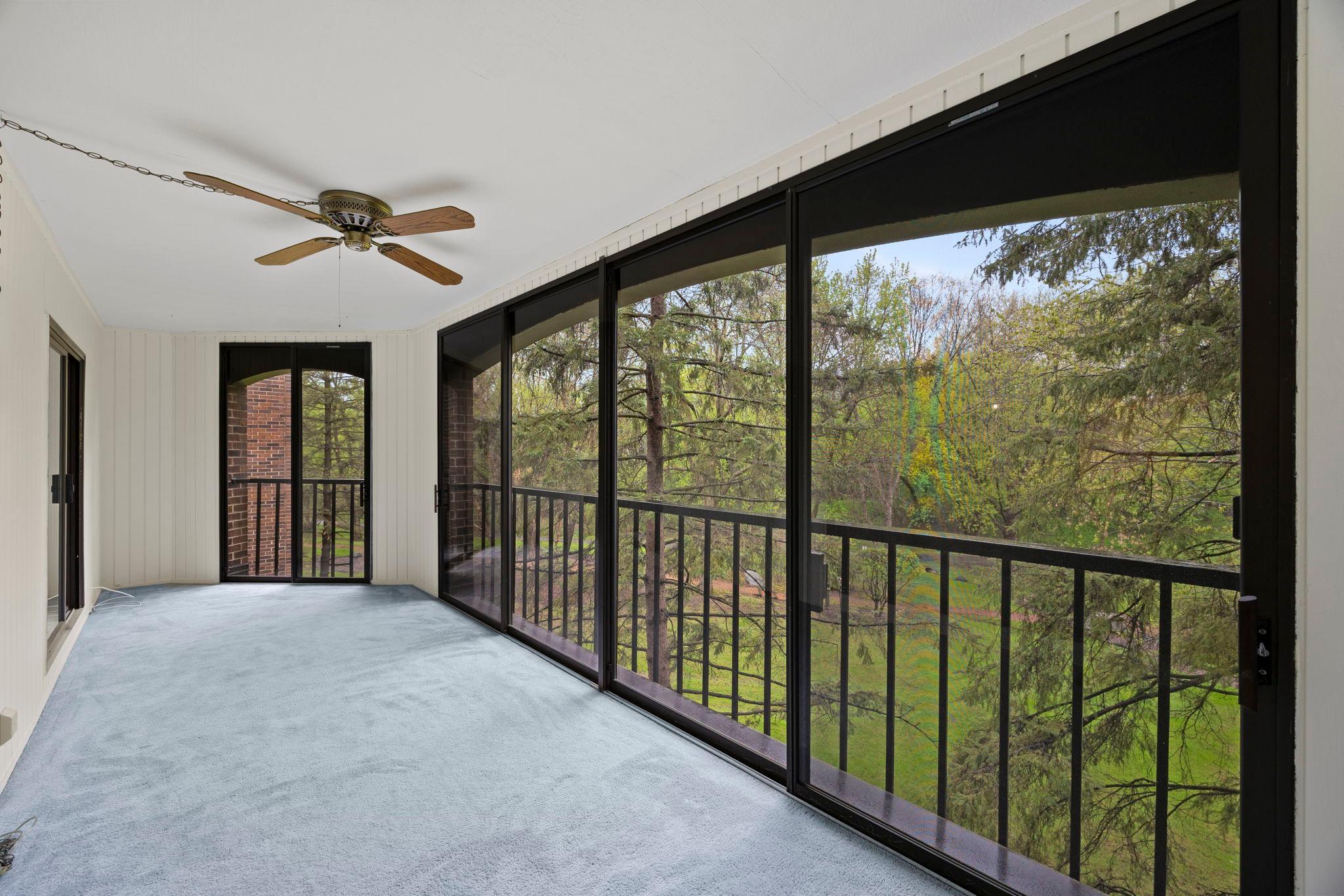 From here, large glass doors extend the living space into an inviting three-season porch, featuring walls of windows overlooking serene green space, cozy carpeting, wood-paneled walls, and an airy, light-filled atmosphere.