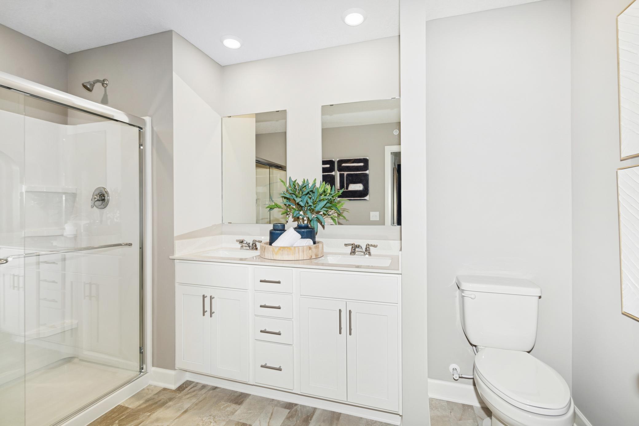 (Photo of model home, features will vary) Featuring a glass-enclosed shower with built-in shelving and glass mirrors set atop dual quartz sinks and vanity-cabinetry, the en-suite bathroom helps streamline morning routines