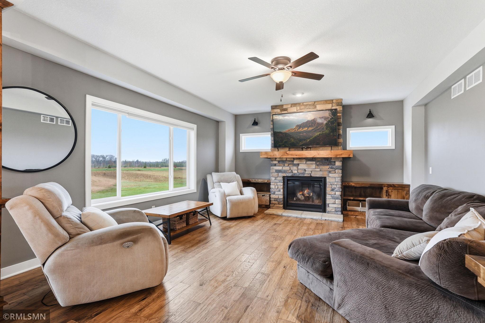 Spacious living room with hardwood floors, a stunning stone fireplace, and large windows that fill the space with natural light and showcase peaceful backyard views. Perfect for relaxing or gathering with family and friends.