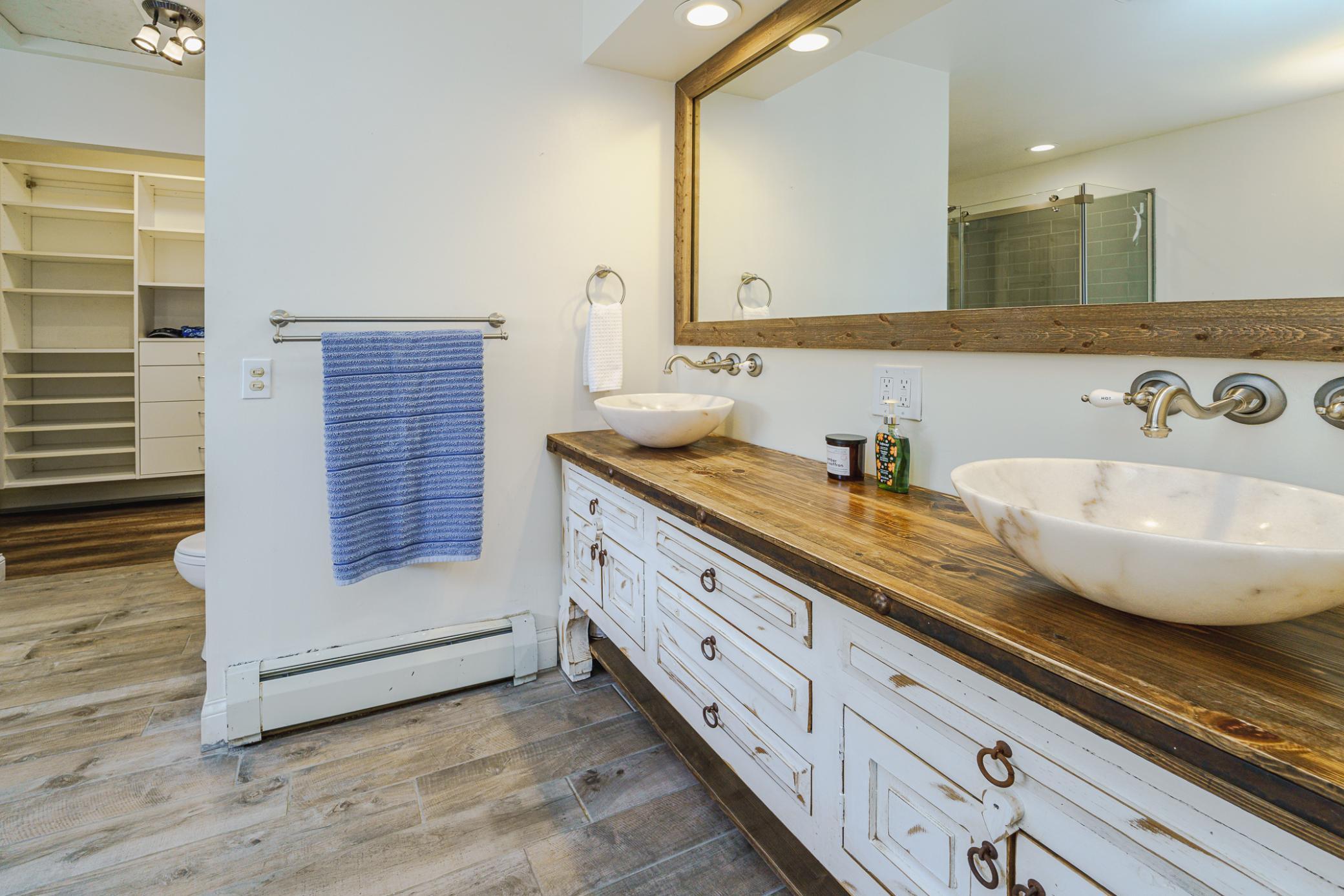 The attached primary bath has plank tile, a furniture-style vanity with dual vessel bowl sinks, a framed mirror, and neutral paint.