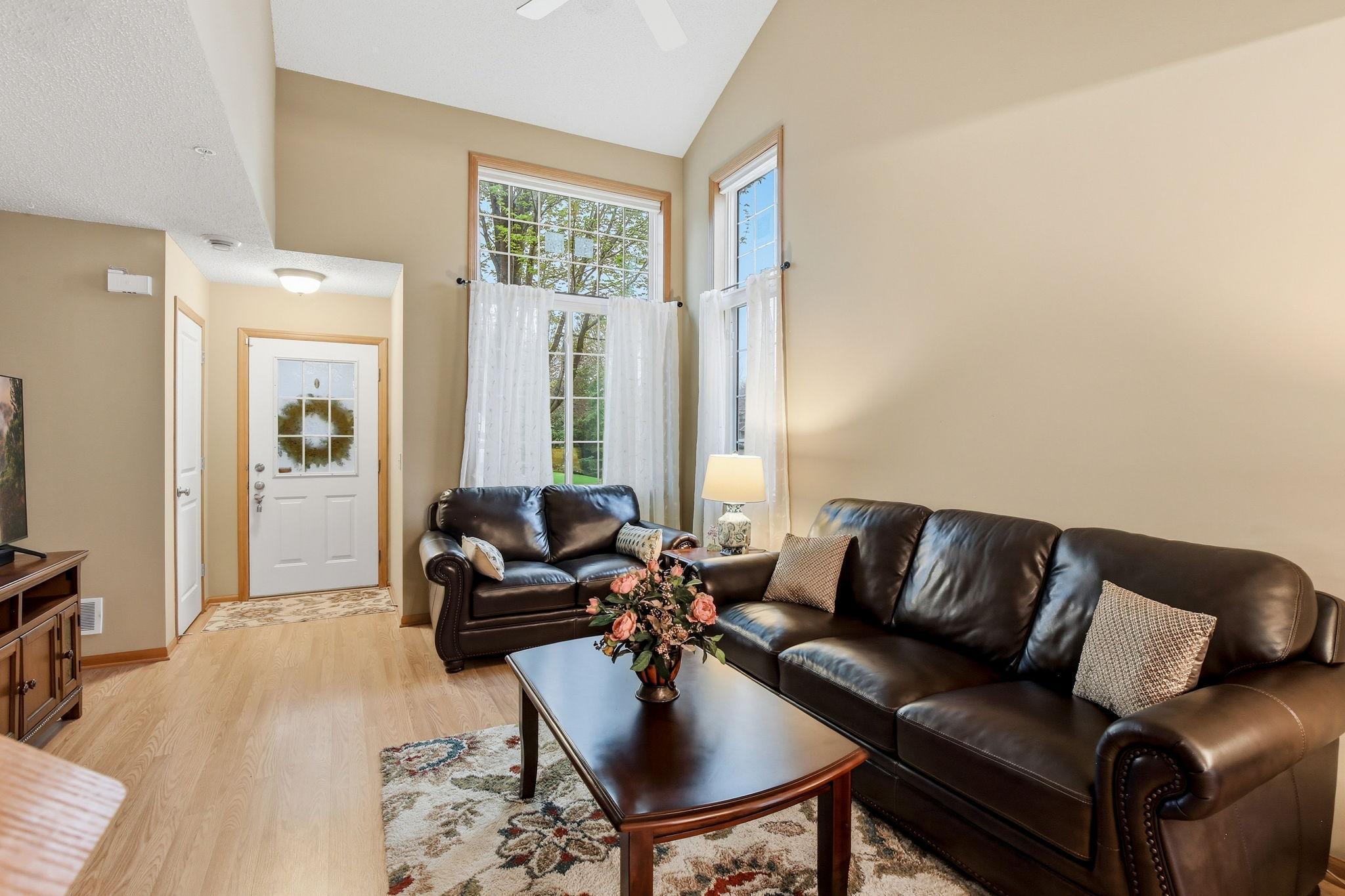 The entry opens into a bright living room with a vaulted ceiling, featuring a convenient coat closet, a ceiling fan, and two large corner windows with remote blinds that fill the space with natural light.