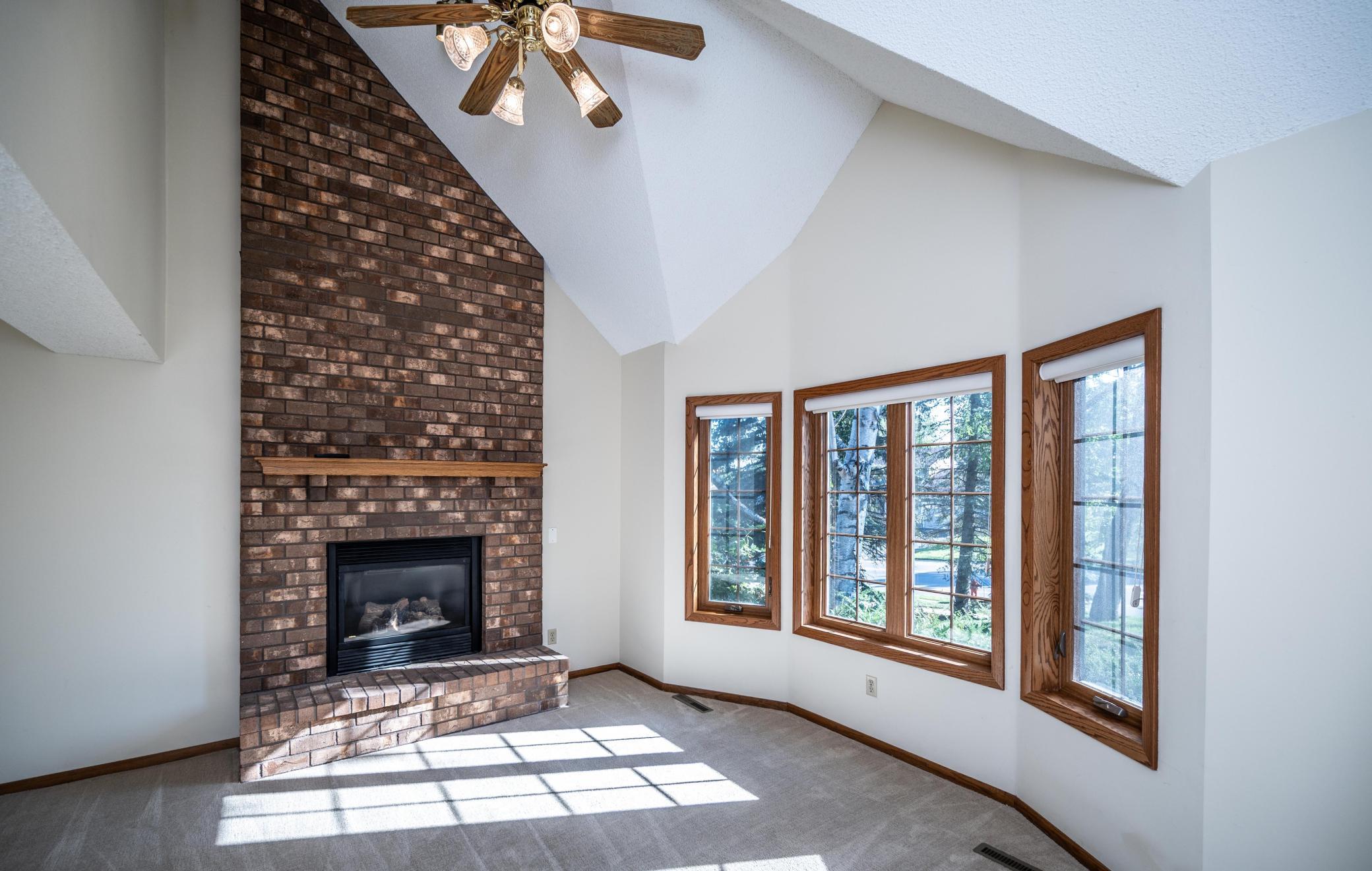 Living room features a gas fireplace with floor to ceiling brick surround, ceiling fan, vaulted ceiling, newer carpet and a bay window.