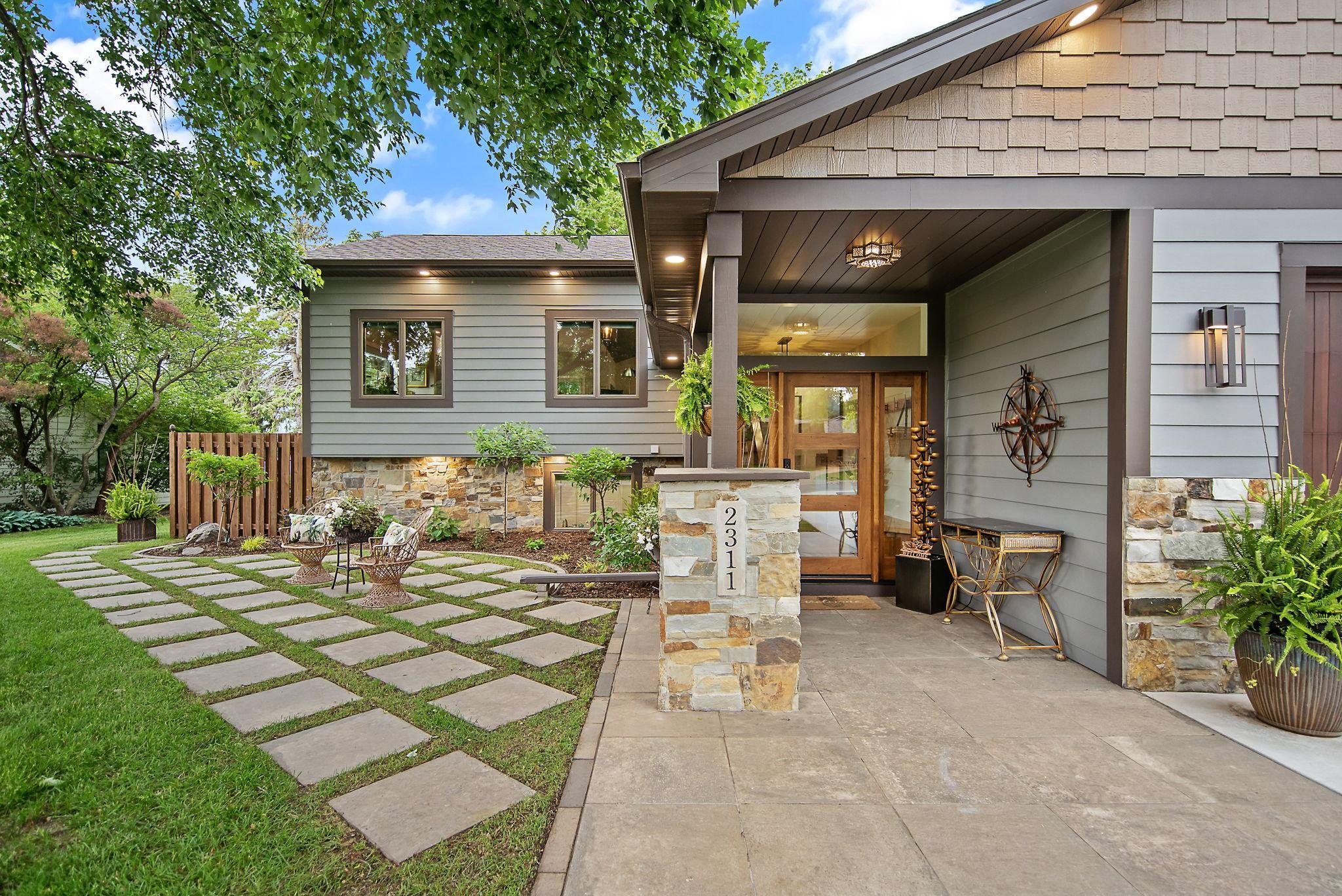 A covered front porch features a porcelain paver entrance and an adjacent green-woven patio makes a warm, inviting entry. A calm, peaceful place to relax and enjoy the scent and beauty of the huge flowering tree in front.