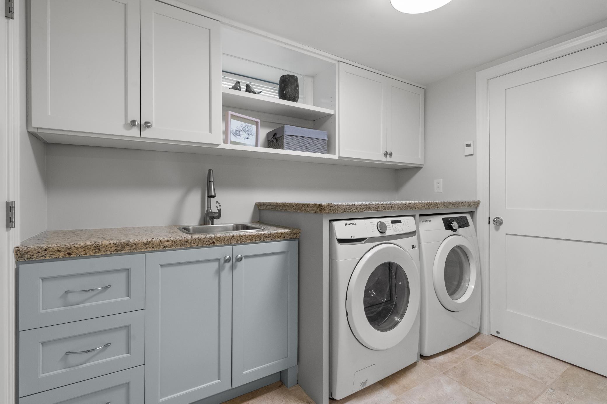 The space features heated floors, walk-in tiled shower, single vanity, front-loading Samsung washer and dryer, and ample granite countertop and cabinetry space with a sink for added convenience.