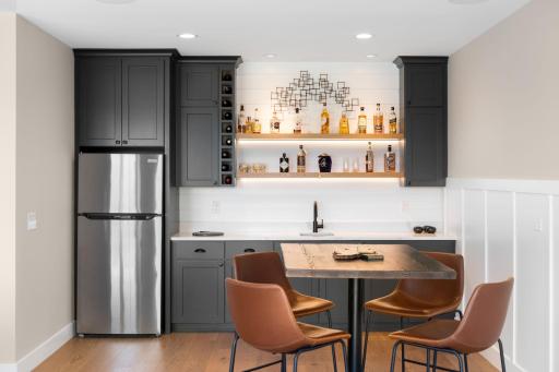 This inviting space flows seamlessly into the bar and entertaining area, complete with custom Iron Ore cabinetry, quartz countertops, a full-size refrigerator, wine rack, open shelving, and a stylish shiplap backsplash.