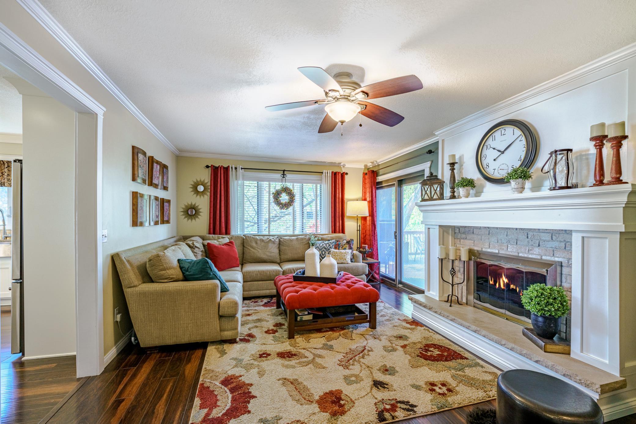 The living room has a substantial fireplace with a dramatic white mantle and brick surround. This room has a ceiling fan and hardwood floors, and it leads to the deck or the kitchen.