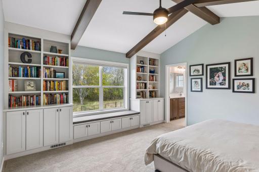 Spacious primary suite featuring vaulted ceilings with wood beams, custom built-ins, ceiling fan, and large window overlooking the wooded backyard.