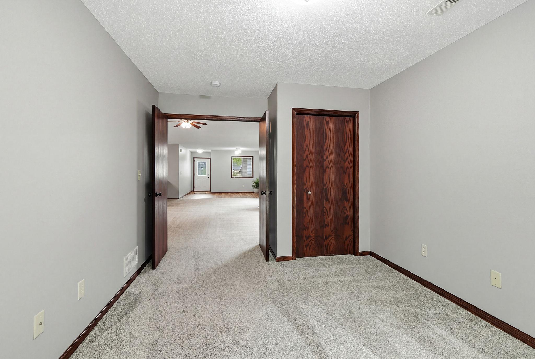 Comfortable carpeted bedroom with neutral walls, rich wood trim, and double doors opening to a bright adjoining living area.