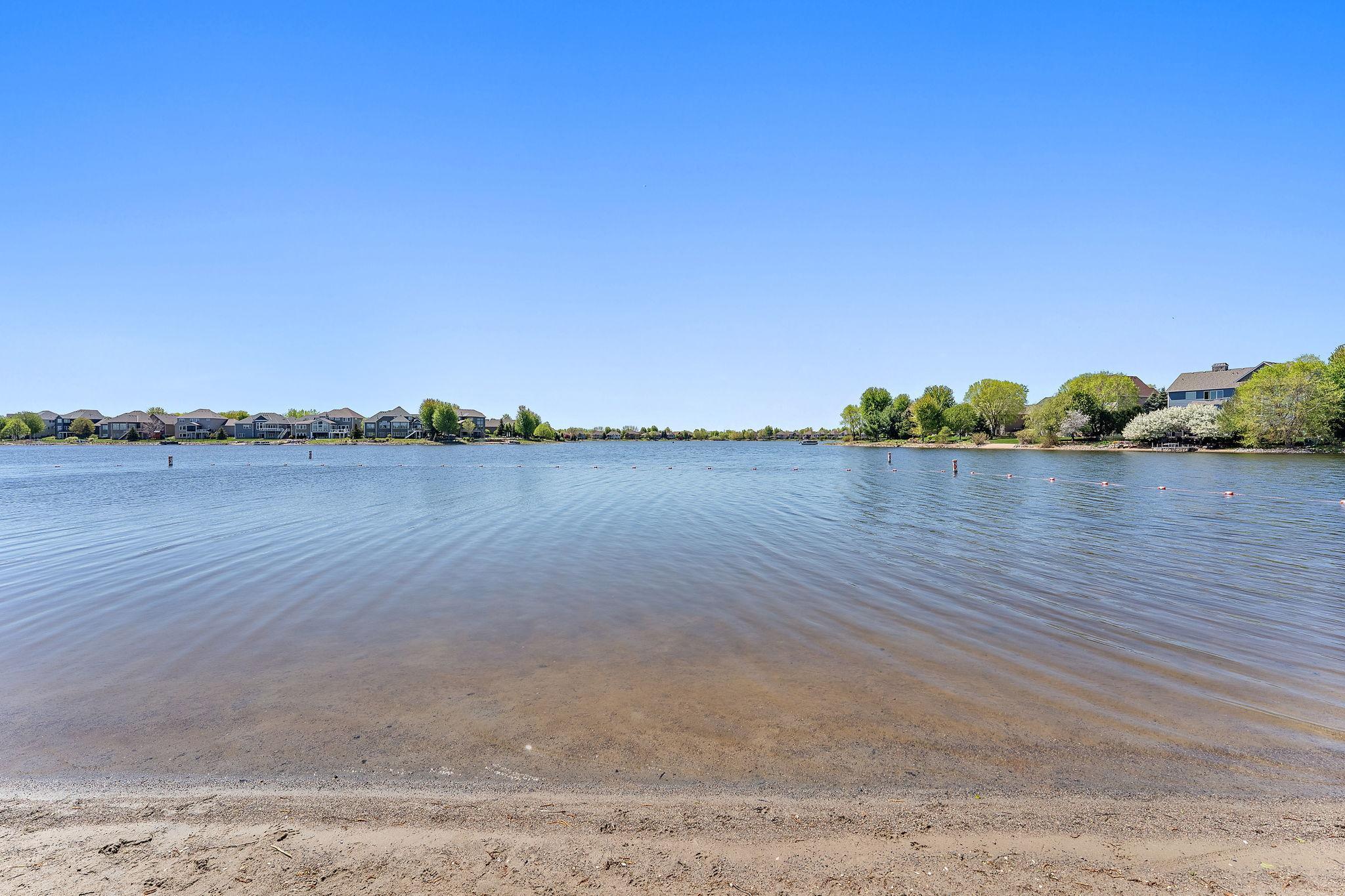 A wide, clean stretch of sandy beach leads into the calm waters of the lake. The water is clear enough near the shore to see the sandy bottom, and the swimming area is neatly marked with buoys