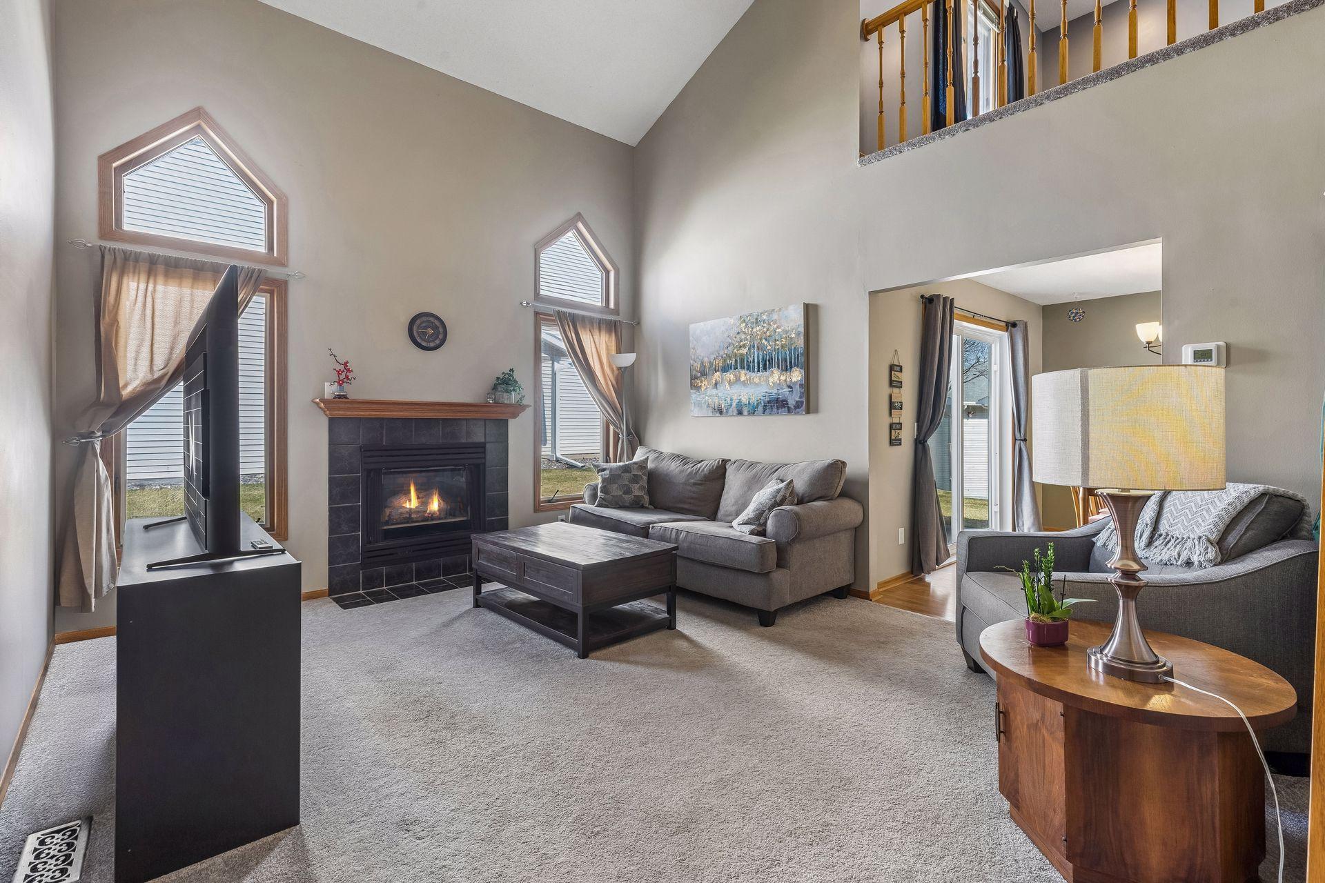 Vaulted living room with abundant natural light from two large windows, featuring plush carpet, a cozy gas fireplace, and ceiling fan with lighting.