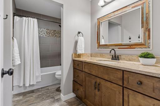 Stylish lower-level bathroom with custom wood vanity, solid-surface countertop, tile flooring, and tile-surround tub/shower combination. Functional and beautifully finished guest bathroom.