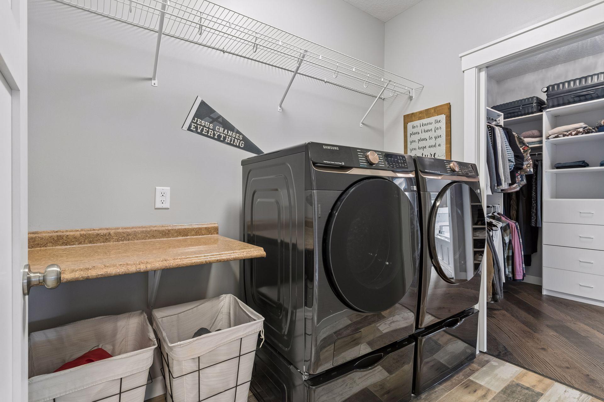 Main-level laundry room with modern washer and dryer, utility counter, extra storage space, and convenient access to the primary suite. Designed for easy one-level living.