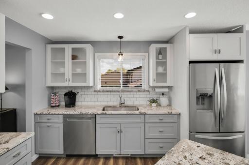 The space features light gray shaker-style lower cabinets paired with crisp white upper cabinets, including elegant glass-front display doors on both sides of the window. Stainless steel appliances, including a French-door refrigerator and dishwasher