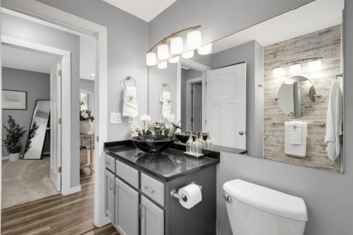 Soft gray walls and white cabinetry provide a clean, neutral backdrop, while the textured accent wall behind the mirror adds warmth & visual interest. The large mirror & modern vanity lighting brighten the room and enhance the open feel of this bath