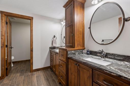 Designed with the same elevated craftsmanship found throughout the home, this stunning lower-level bath features dual vanities, rich custom cabinetry, designer tile floors, and a private shower and water closet, offering comfort and sophistication.
