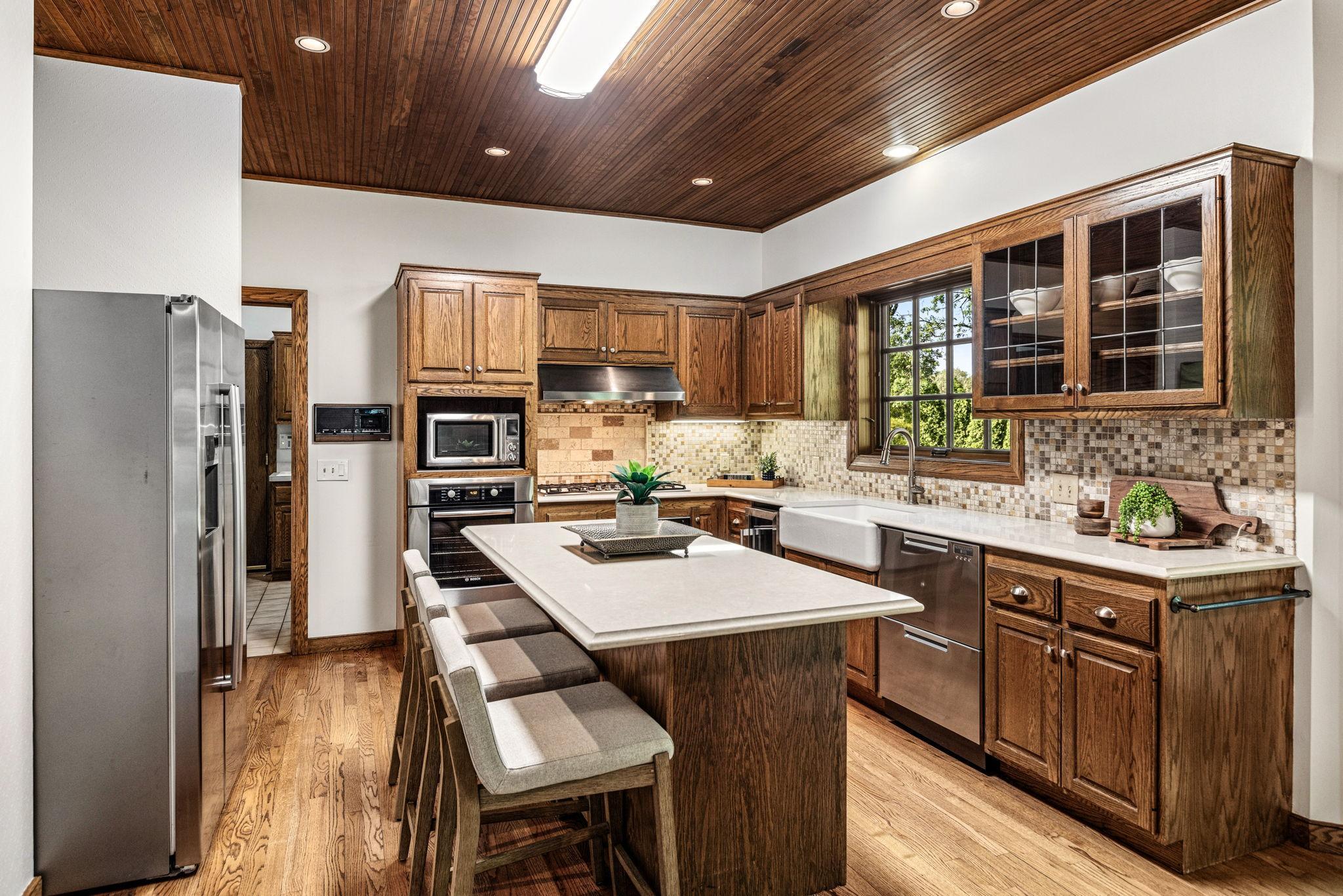 Warm and inviting kitchen featuring rich wood cabinetry, hardwood floors and a center island with seating. Stunning tongue-and-groove wood ceiling and expansive window brings warmth and natural light to the heart of the home.