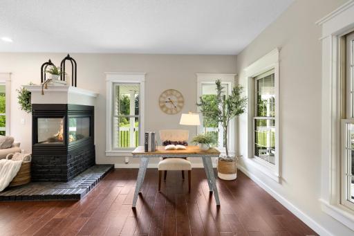 Versatile living room or home office featuring rich hardwood floors, abundant natural light, and a dramatic two-sided gas fireplace shared with the adjoining family room.