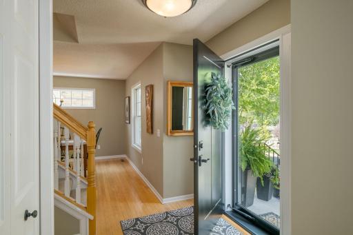 The entry sets the tone: warm hardwood floors, a graceful staircase with classic white balusters, and a glass door that frames the lush greenery beyond like a living painting.