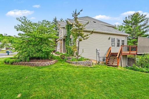 This side view showcases the lush gardens and direct deck access from the stairs. Highlights include durable steel siding and stone accents, a concrete driveway, and a finished two-car garage with a service door, window, and two openers (2024).