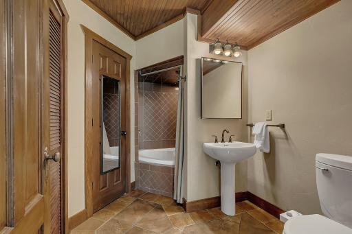 Lower-level bathroom boasting a jetted tub with tile surround, a handsome pedestal sink, a built-in linen closet, and an adjacent laundry closet for maximum efficiency.