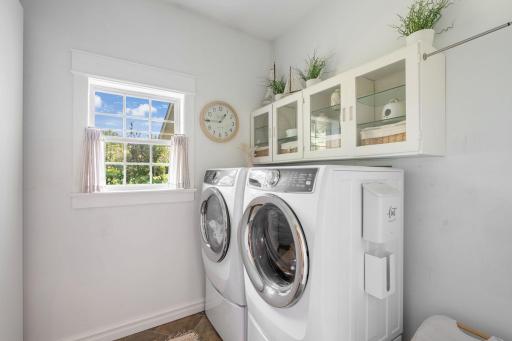 Nearby, the main-level laundry room offers a front-loading washer and dryer, overhead glass-front cabinetry, and abundant storage.