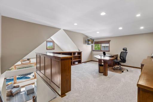 The upper level features a private office space. It includes newer carpet (2021), a wall of built-in shelves, cabinets & a large built-in desk. The room is lit with recessed lights & natural light from the window.