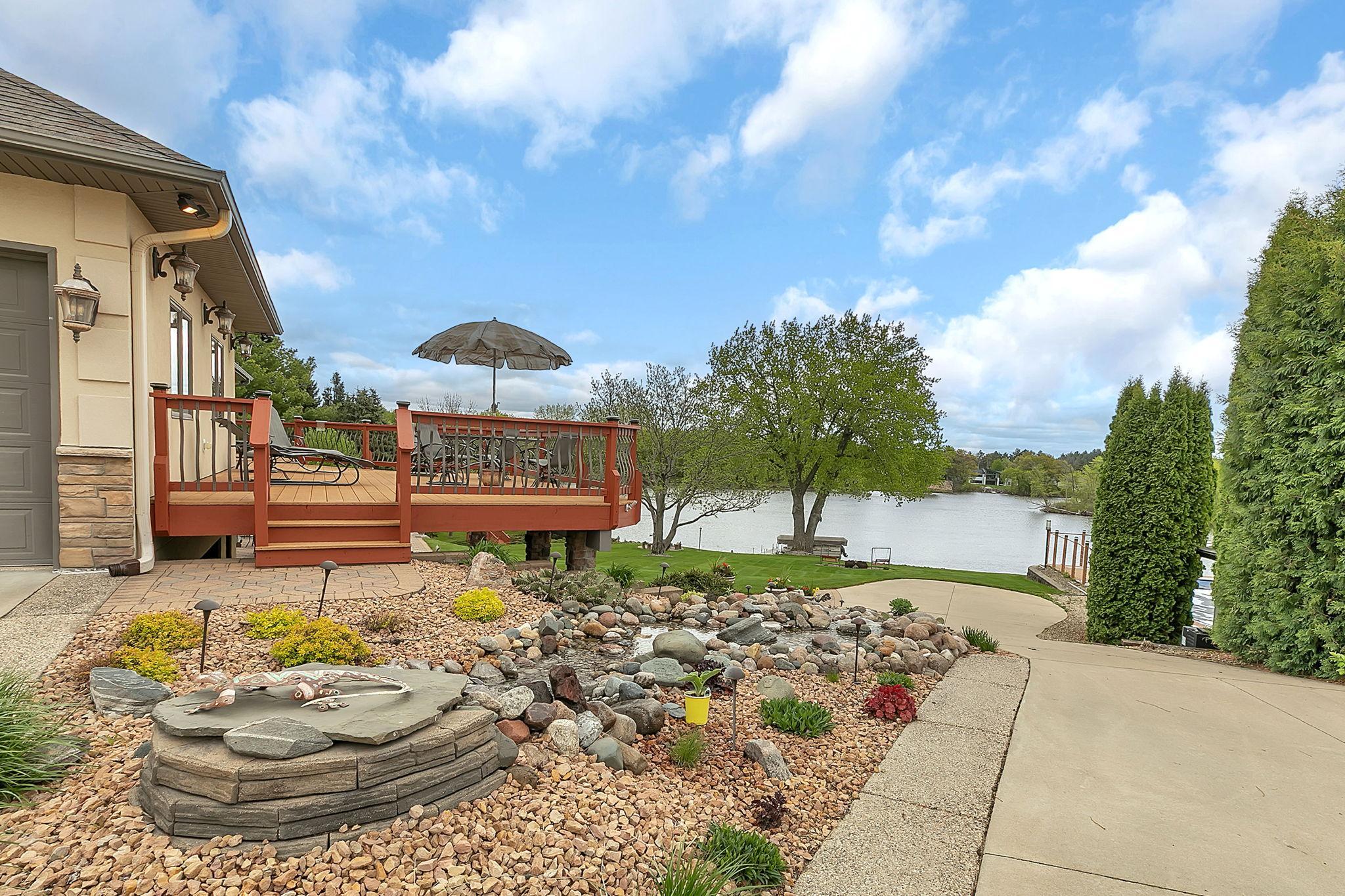 Serene waterfall winds through the landscape, creating a tranquil setting. Deck space wraps from slider at the dining room around the garage. To concrete on the right is the driveway access to the lower tuck under garage.