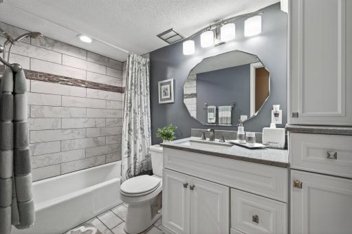 Updated upper-level full bathroom with marble-look tile surround on the tub/shower, white cabinetry, updated vanity, modern geometric mirror, and new lighting.