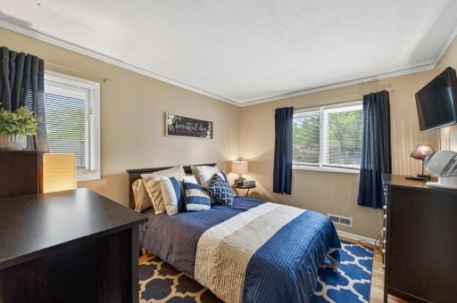 Upper-level bedroom with hardwood floors, two windows with tree views, and warm neutral tones â a comfortable, well-proportioned space.