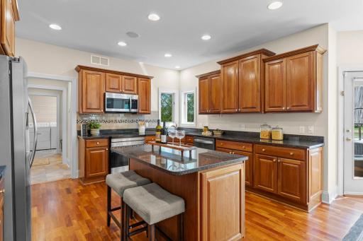 The kitchen is a dream, showcasing abundant wood cabinetry, expansive granite countertops, a center island with breakfast bar seating, stainless steel appliances, recessed lighting, and hardwood flooring.