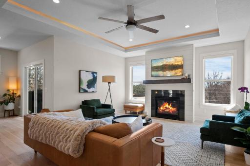 A cozy living room is accented by a gas fireplace, in-ceiling tray lighting, ceiling fan and views of the backyard