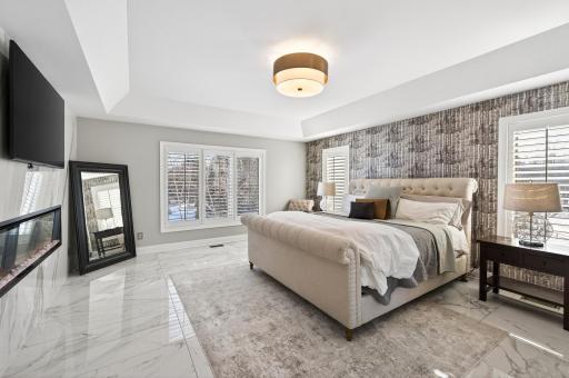 Enter through double doors into a serene retreat featuring tray ceilings, marble flooring, and a cozy electric wall fireplace with a marble surround. Large windows provide abundant natural light, while two generous walk-in closets with built-ins.