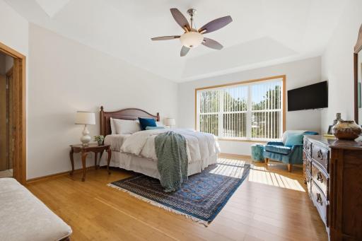 The spacious primary bedroom is conveniently located on the main floor & features a tray ceiling, abundant natural light, & natural wood flooring. The room easily accommodates a king-size bed as seen here, highlighting its generous proportions.