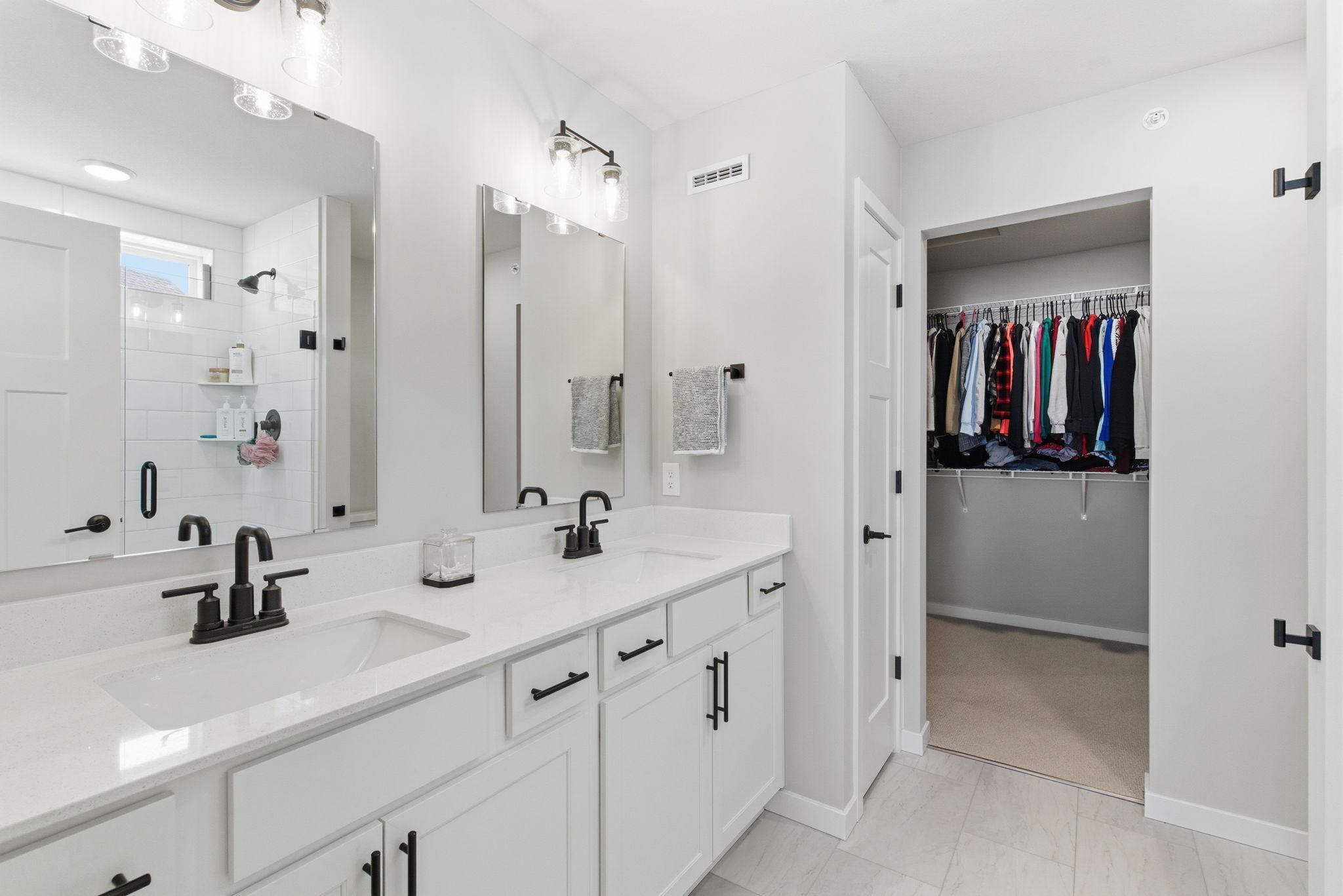 A refined primary ensuite offers spa-inspired comfort and modern sophistication. The bathroom features a sleek double vanity with quartz countertops, contemporary black fixtures, and oversized mirrors illuminated by modern wall sconces.