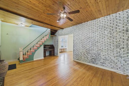 Inviting living room with warm wood-look flooring, a striking painted brick accent wall, and beautiful wood plank ceilings that add character and charm. The spacious layout offers plenty of room for gathering and opens nicely adjacent room.