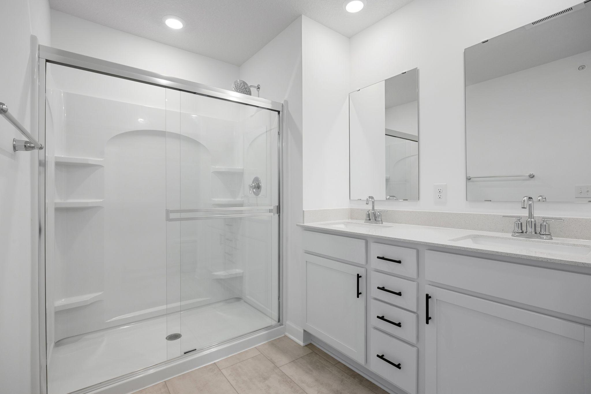 Featuring a glass-enclosed shower with built-in shelving and glass mirrors set atop dual quartz sinks and vanity-cabinetry, the en-suite bathroom helps streamline morning routines