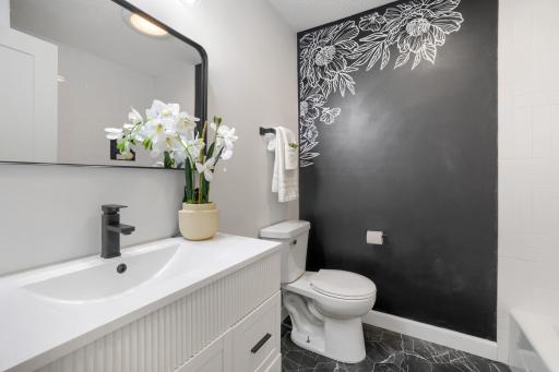 This beautifully remodeled hall bathroom showcases a sleek white vanity with contemporary detailing is paired with a quartz countertop, updated black fixtures, & a stylish framed mirror, black tile floors creating a clean & elegant aesthetic design.