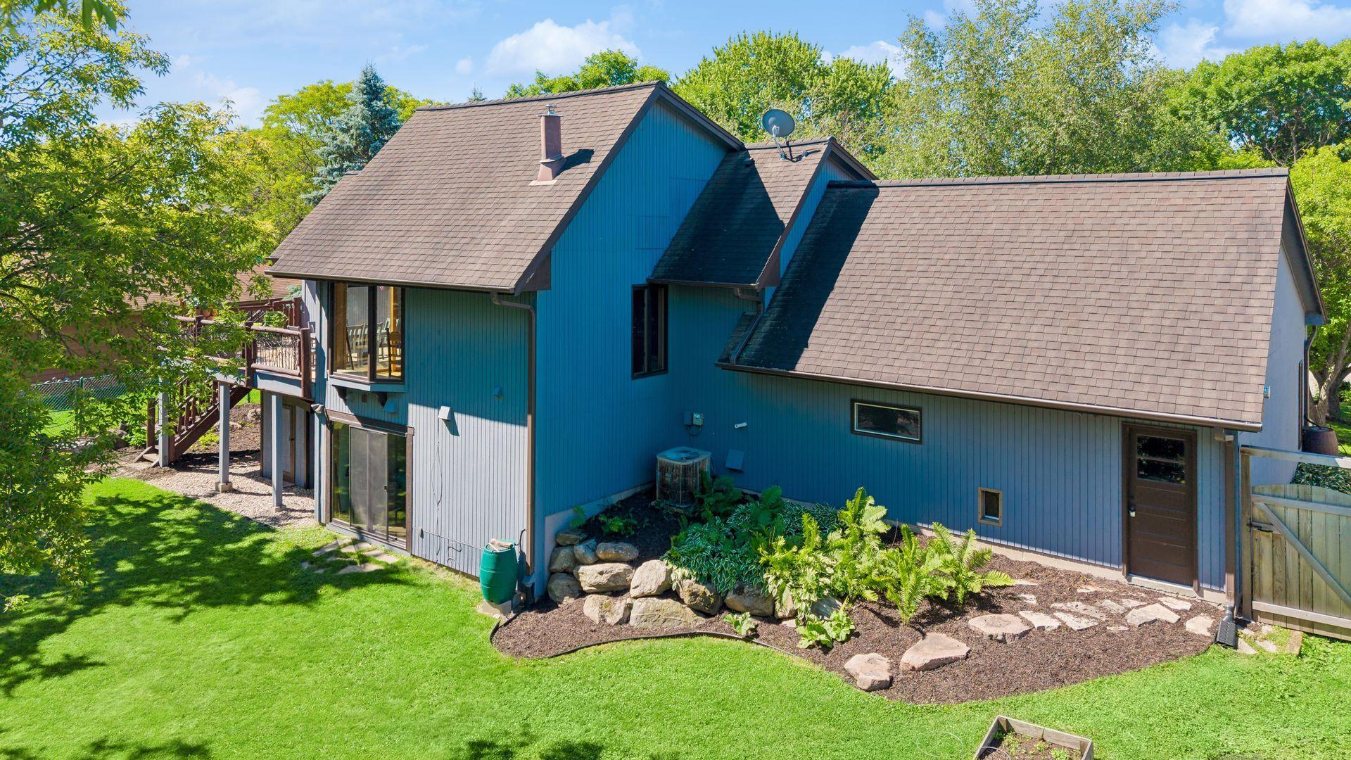 Rear view of home with lovely perennial landscaping, rain barrels, three sets of sliding patio doors, attached two-car garage and deck overlooking the lake.